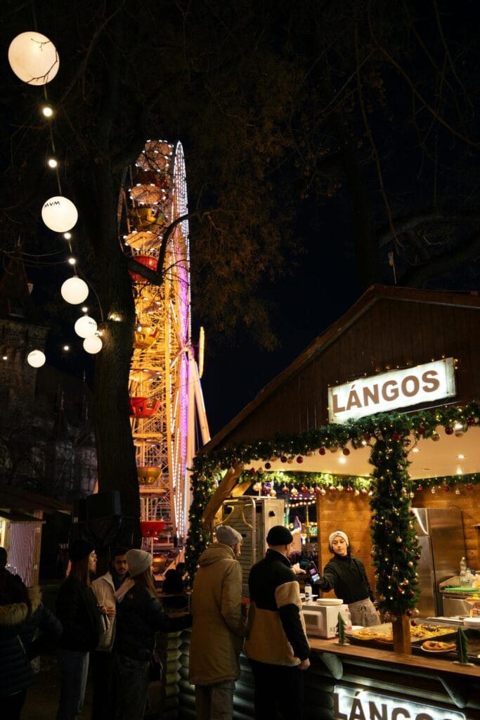Puesto de lángos en un mercado navideño de Budapest con una noria iluminada al fondo durante la noche.